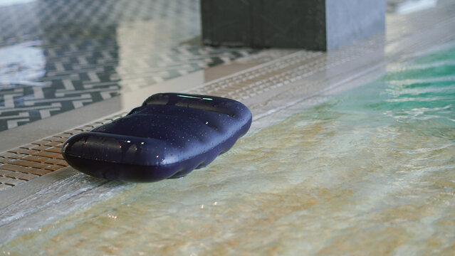 A dark blue inflatable pool pillow or float rests on the wet edge of an indoor swimming pool next to a patterned tiled floor