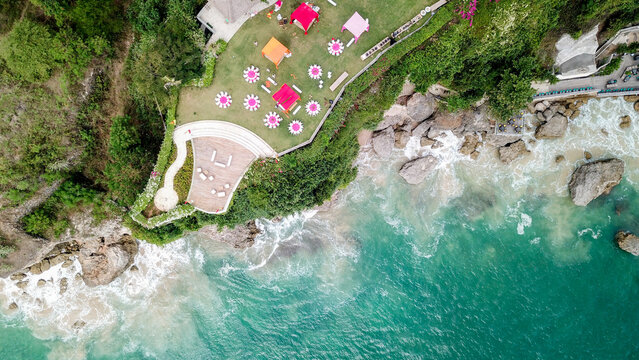 Indonesia - 04 July 2018: Aerial view of an outdoor event setup on a grassy cliffside overlooking the turquoise ocean with colorful tents, circular tables, and a wooden deck.