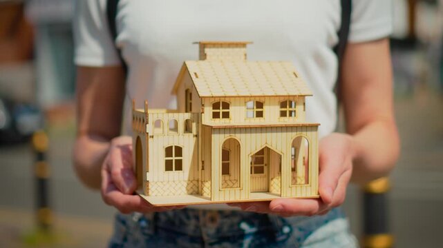 Woman holding wooden house model in hands. Property purchase and mortgage loan concept with miniature home and real estate investment planning. Residential ownership and moving idea with suburban