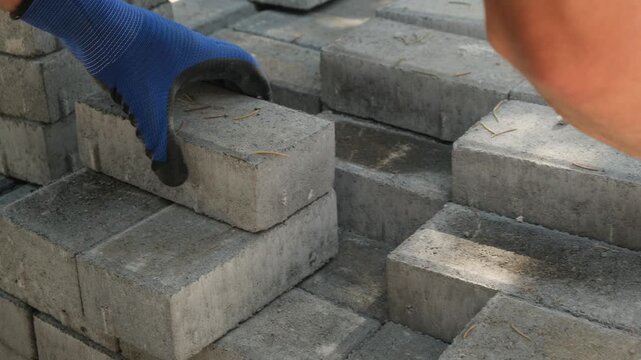 Construction worker in blue gloves prepares a small stack of gray paving tiles for the next worker. Material handling, construction teamwork, municipal work and migrant workforce