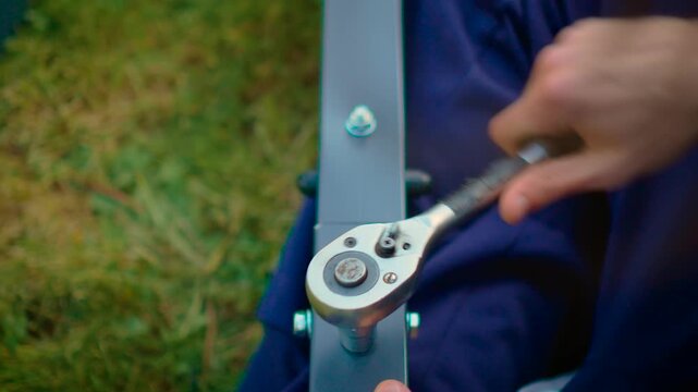 Hands rotating socket wrench securing fastener on steel frame element in daylight. Mechanical assembly process with equipment and durable materials in garden environment. Industrial maintenance