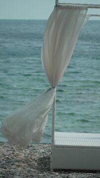 Beach, ocean, canopy, white fabric blowing in gentle breeze on tranquil pebble beach, vacation, resort. Vertical video