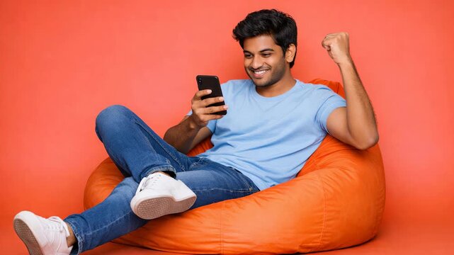 Young man relaxes on orange bean bag chair. He holds phone while smiling and flexing his right arm. Bright orange background emphasizes casual, cheerful mood