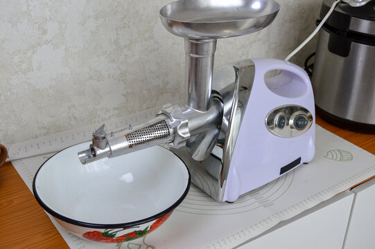 Electric meat grinder with juicer squeezer attachment standing near enamel bowl on kitchen table. Appliance for making tomato juice, food prep equipment, extract attachment and domestic cooking concep