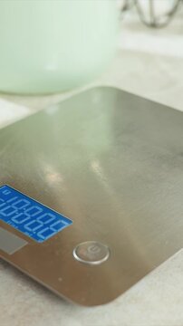 Hand presses power button on digital kitchen scale. Metallic surface reflects light under close up view. Electronic weighing machine has blue LED display and brown casing