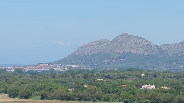 Wide shot of a rural countryside landscape near Begur, Spain, featuring green fields, scattered houses, and a prominent rocky mountain range under a clear blue sky.