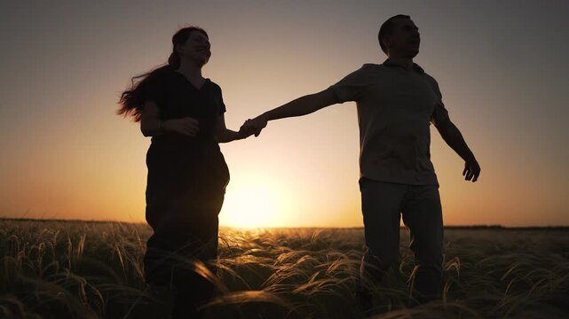 Happy couple holds hands walking through tall grass field at sunset. Man and woman silhouettes move against golden sky. Couple enjoys romantic walk outdoors. Sun glows on horizon behind them.