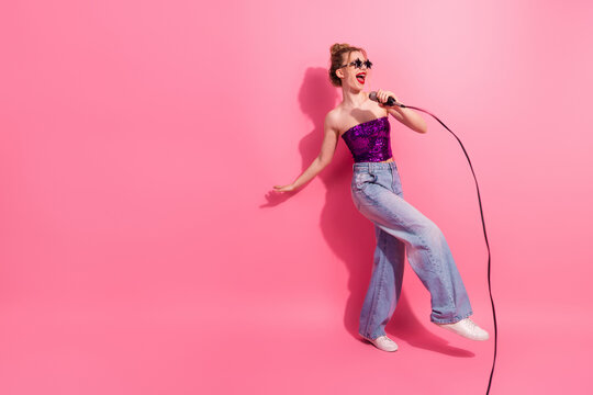 Cheerful stylish woman singing into a microphone wearing glittery attire on a vibrant pink background, expressing joy