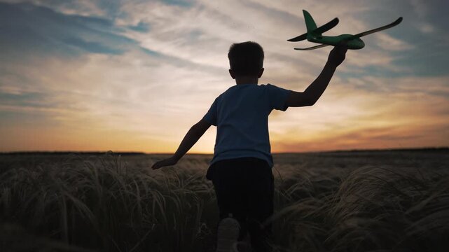 Happy boy runs through field at sunset holding toy airplane. Child plays with plane in grass during golden hour. Kid dreams of flight while running outdoors. Boy silhouette runs freely with airplane.