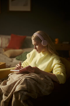 Young Caucasian woman wearing headphones and writing in notebook while sitting in armchair. Image represents remote learning and mental wellbeing, vertical