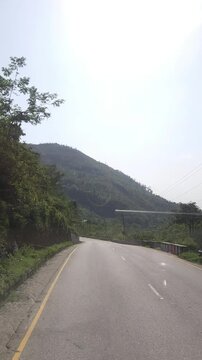 Vertical footage of a scenic paved highway winding through lush green mountain terrain in Nepal. The road passes between densely vegetated hills with utility lines and roadside safety barriers