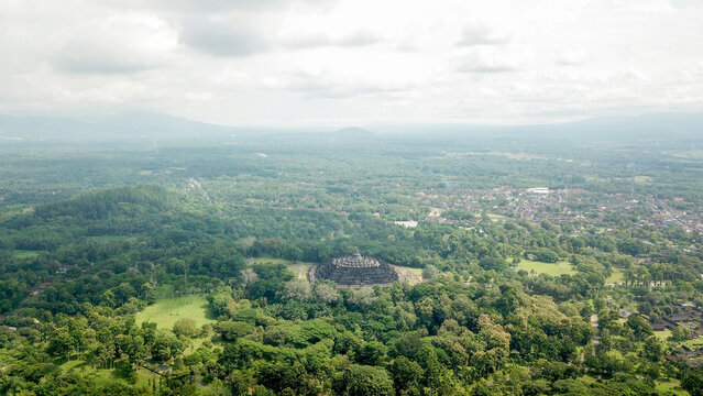 Indonesia - 23 February 2023: Aerial view of Borobudur Temple surrounded by lush green forests and distant mountains under a cloudy sky.