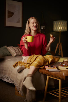 Vertical image of happy Caucasian young woman sitting on bed, holding mug and eating snack. Scene represents morning routine, self care, and cozy lifestyle marketing campaigns