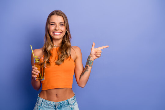 Young chic woman in orange crop top and denim shorts holding a drink smiles and points against a purple background