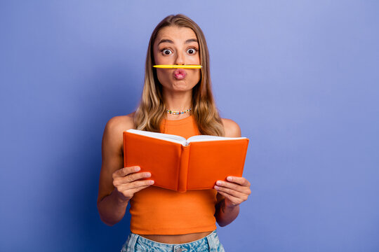 Young chic student reads orange notebook with pencil balanced on lips against blue background