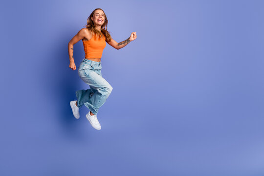 Young chic woman jumps in orange singlet and jeans against purple background showcasing energy joy and fashion
