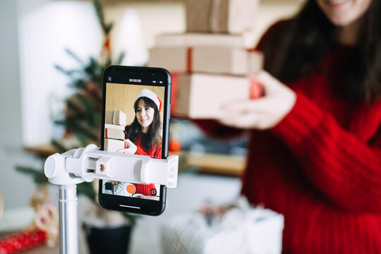 Phone on tripod films woman in Santa hat holding stack of wrapped parcels at home. Influencer marketing holiday season, small business video promotion, content creator business marketing