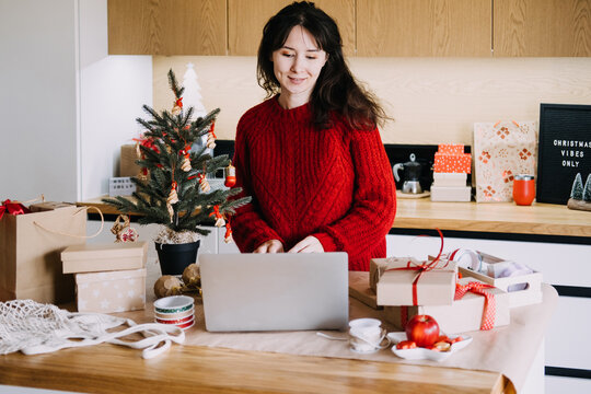 Woman in knitted sweater works on laptop at kitchen table beside decorated tree and wrapped gifts. Balancing work christmas season, holiday time management concept, juggling work and holidays