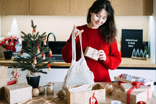 Woman holds mesh bag and kraft wrapped parcel in kitchen near CHRISTMAS VIBES ONLY sign. Eco christmas gift business, sustainable gift shop christmas, handcrafted christmas gift boxes business