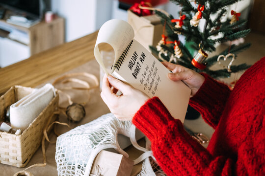 Hands hold notebook with HOLIDAY BUDGET PLAN beside wrapped gifts and small Christmas tree. Holiday budget planning, mindful christmas spending, no impulse buying gift rules