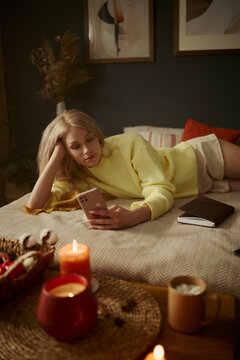 Vertical image of young Caucasian woman lying on bed and using smartphone. Image represents digital life and relaxation, suitable for mental wellbeing and lifestyle marketing