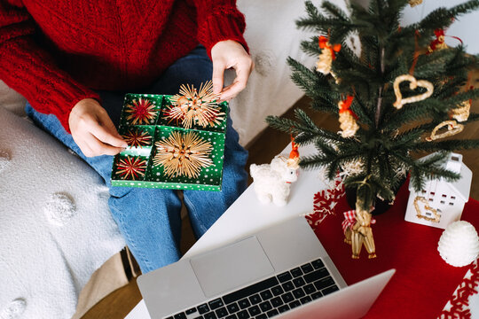 Woman holds box with straw star ornaments on lap beside laptop and small decorated tree. Inherited christmas ornaments, family holiday heirloom, grandmother decorations box