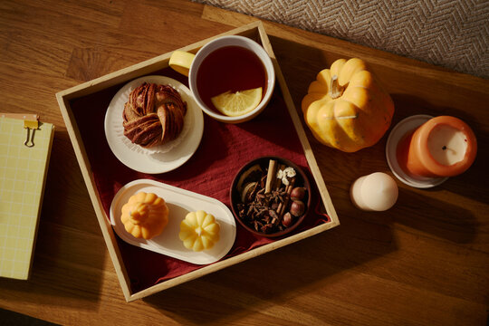 Wooden tray holding hot tea with lemon, cinnamon roll, spices, and pumpkin candles on table. High angle shot representing autumn lifestyle, suitable for seasonal marketing campaigns