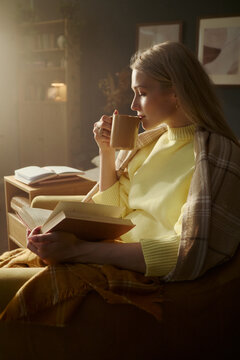 Young Caucasian woman sitting in armchair, reading book and drinking hot beverage, representing mental wellbeing and self care. Image suitable for lifestyle marketing, vertical