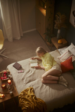 Vertical image of young Caucasian woman lying on bed and reading book. High angle view of female student studying at home, suitable for mental wellbeing and lifestyle marketing