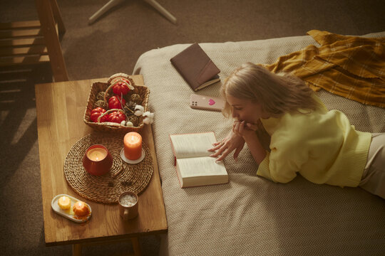 Young Caucasian woman lying on bed and reading book next to table with burning candles. Image represents mental wellbeing and self care, suitable for lifestyle blog, high angle view