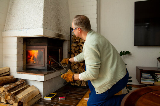 Mature man adding firewood to fireplace at home, heating, energy, cozy winter lifestyle and domestic comfort concept in modern living room
