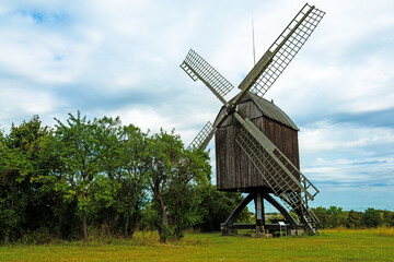 Historic wooden post mill with large sails standing in a green rural landscape under a cloudy summer sky. Horizontal format. © Heidi