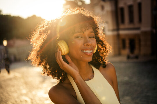 Young woman wearing yellow headphones smiling in golden sunlight outdoors on a city street enjoying music and summer vibes