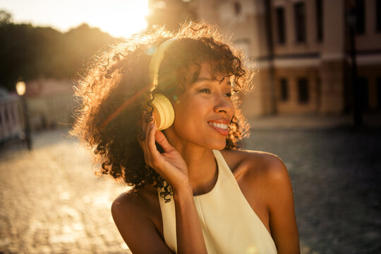 Young woman enjoying music with headphones outdoors at golden hour in a city street, smiling and relaxed while listening to songs