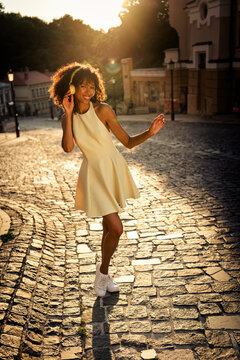 Young woman dancing with headphones in a yellow dress on a cobblestone street at golden hour, smiling and carefree during sunset in an urban historic setting
