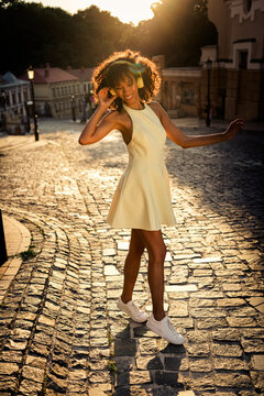 Young woman dancing on a cobblestone street at sunset wearing headphones and a yellow dress, enjoying music and golden hour in the city