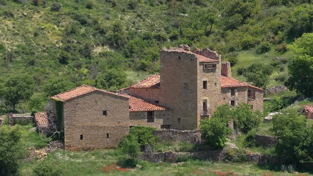 A static wide shot of a historic stone building with a tower and red-tiled roofs, surrounded by lush greenery and low stone walls in the countryside near Mirambel, Spain.