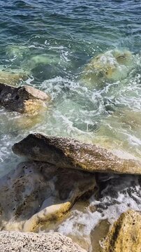 Ocean waves crash against limestone rocks at the Mediterranean coastline on a sunny day. High-angle static close-up shot.