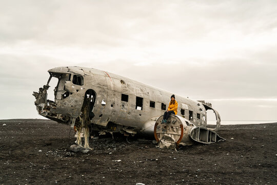 A white woman sits on wreckage of a crashed airplane on a black sand