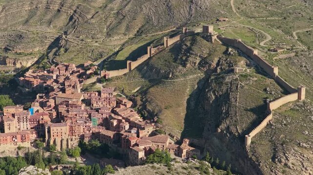 High-resolution aerial footage showcasing the historic town of Albarracin in Spain, featuring its iconic medieval city walls winding over a rugged hilltop. No people are visible in the frame.