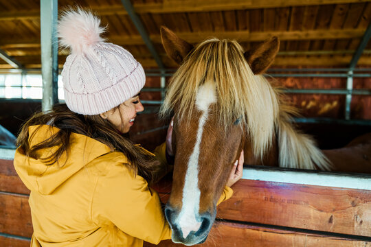 A white woman gently pets a brown horse inside a wooden stable