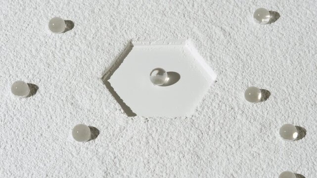 Top-to-bottom curved shot of white sifted layer of powder with hexagonal imprint and hydrogel spheres chaotically laid over the surface lit by rotating light | Moisture retaining effect commercial