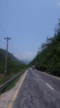 Scenic driving footage of a paved mountain highway winding through lush green forested hills in Nepal. The road passes through a beautiful natural landscape beneath a clear blue sky