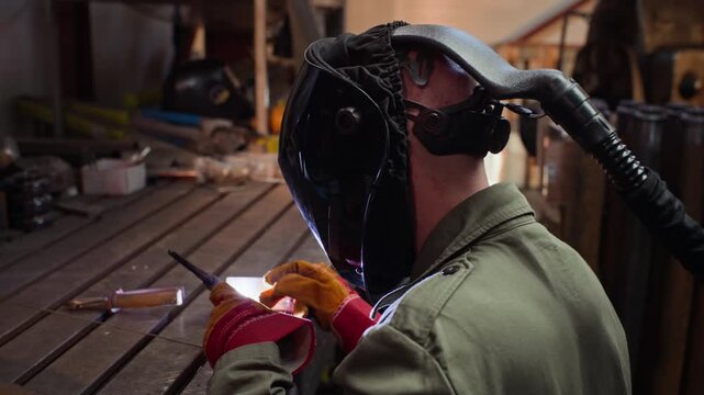 Male welder wearing protective helmet welding metal plate on workbench in workshop. Footage is suitable for industrial manufacturing and vocational training videos