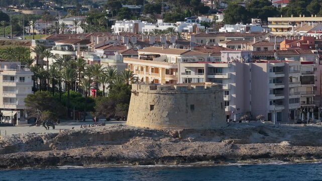 A scenic coastal view of Moraira, Spain, featuring the iconic stone watchtower on rocky cliffs with the Mediterranean Sea in the foreground.