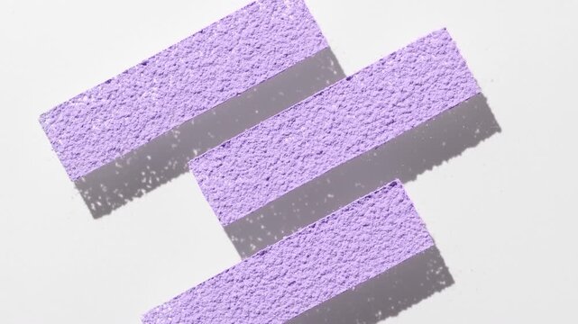 View from above shot of pastel violet powder sifted through three stripe shaped stencil on transparent surface casting moving shadow on light grey background underneath