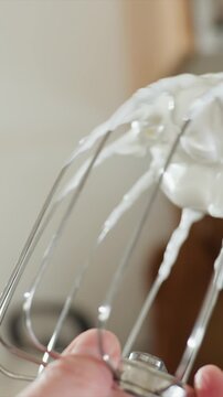 Close up of hand holding electric whisk attachment covered. Creamy texture forms peaks and drips around metal whisks as they are. Blurred kitchen background suggests indoor setting