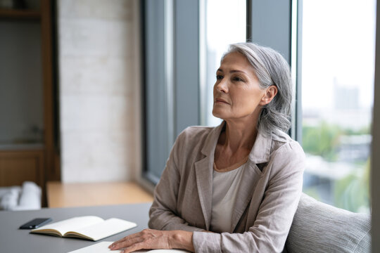 A thoughtful older woman sitting in a modern office space, staring out of the window, reflecting on her day and pondering future ideas amidst a minimalist decor.