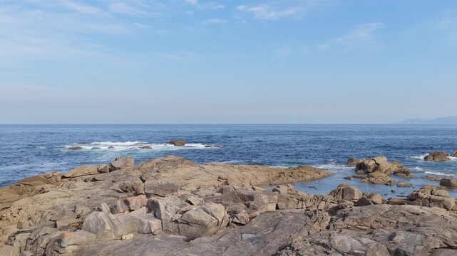Wide shot of a rugged rocky coastline along the Atlantic Ocean in Baiona, Spain. The video shows waves gently breaking over large rocks under a bright blue sky with scattered clouds.