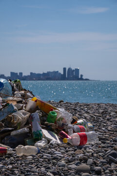 Plastic waste and trash piled up on the pebble beach, with the city in the background, highlighting pollution and the impact of human activities near the city's coastal areas. Vertical photo